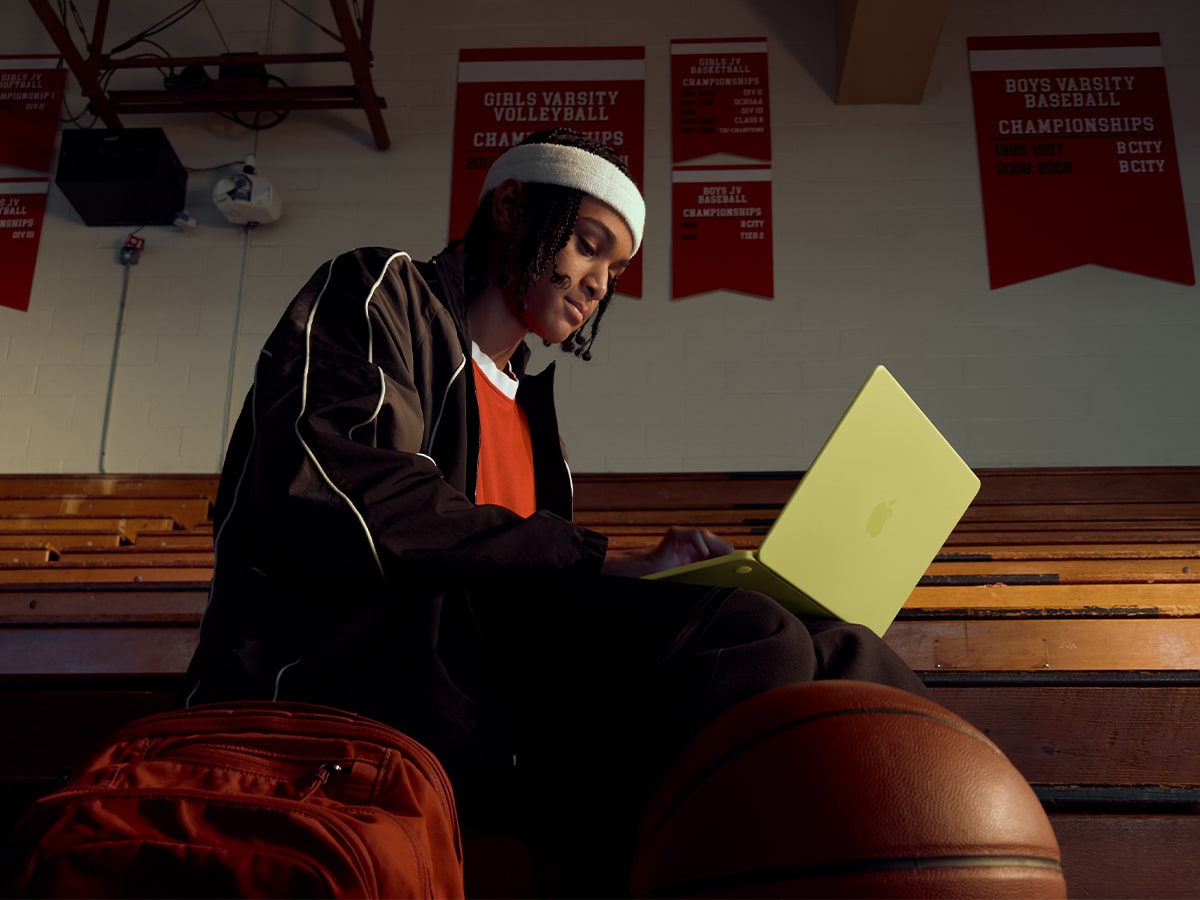Person using yellow MacBook Neo on gym bleachers, wearing a black tracksuit and white beanie, with a basketball nearby.