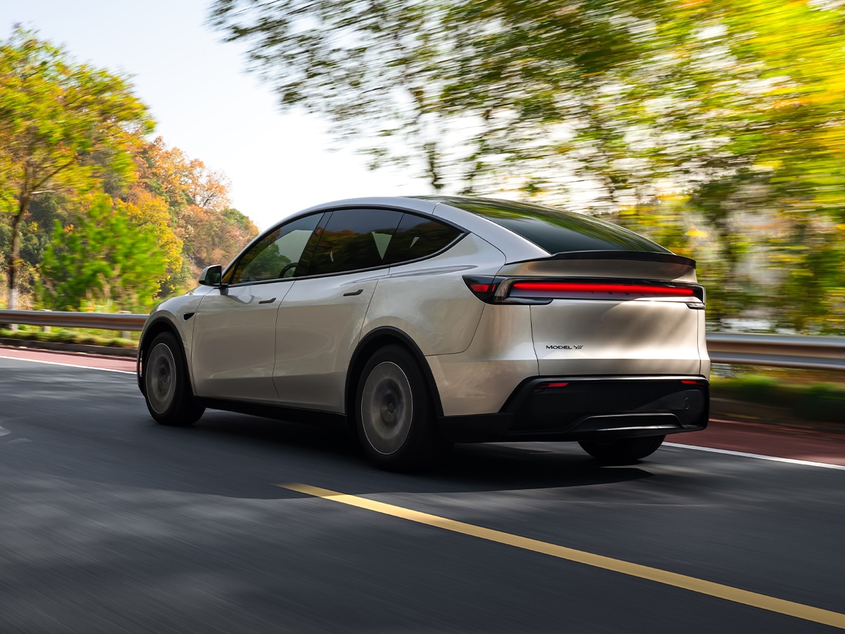 Tesla Model Y L in white driving on a scenic road, rear three-quarter view with blurred greenery.