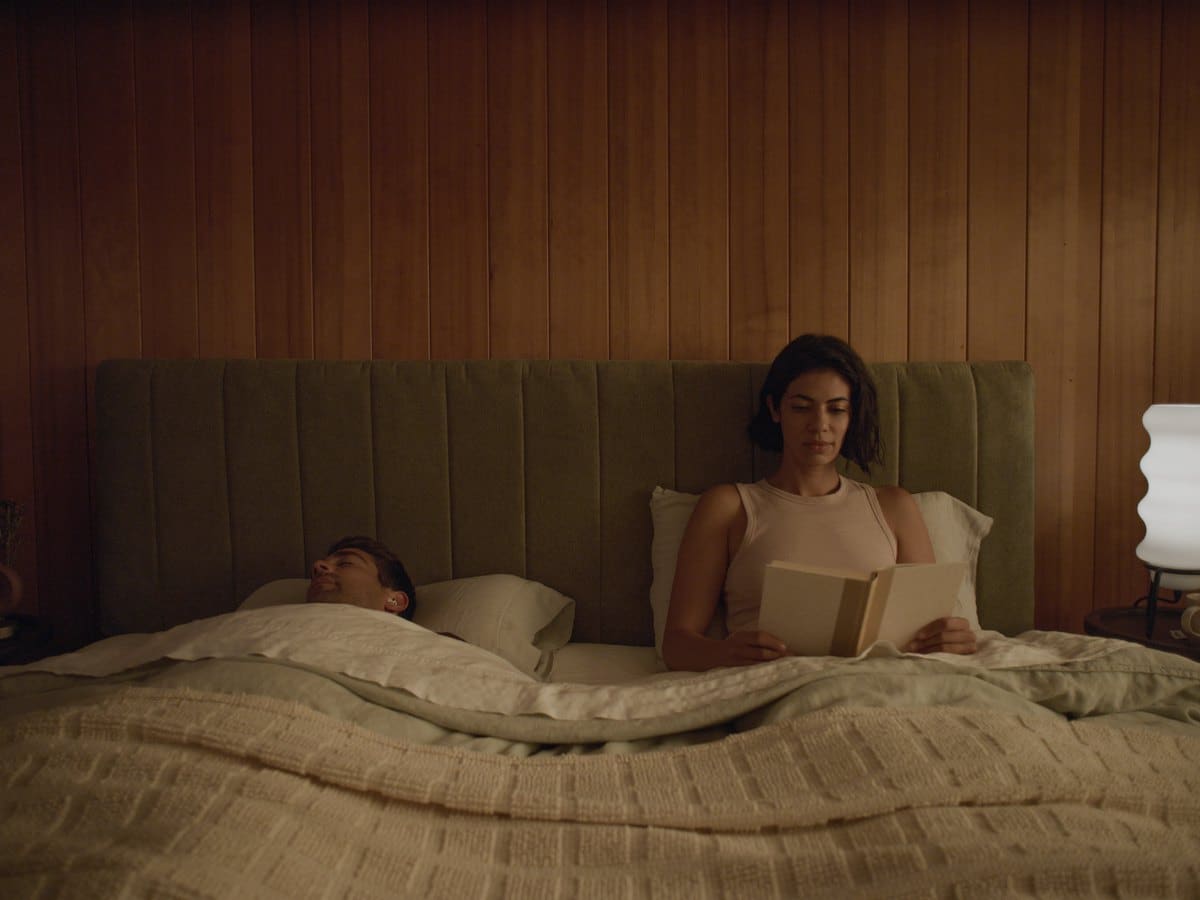Person reading in bed by lamplight beside a partner asleep with Ozlo Sleepbuds