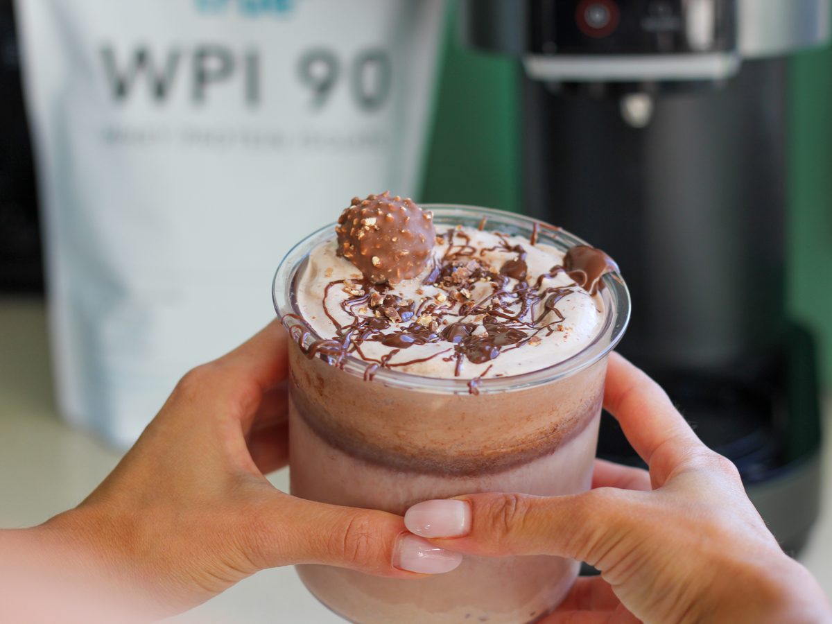Hands holding a Ferrero Rocher protein shake topped with whipped cream and chocolate, with a True Protein WPI 90 pouch behind