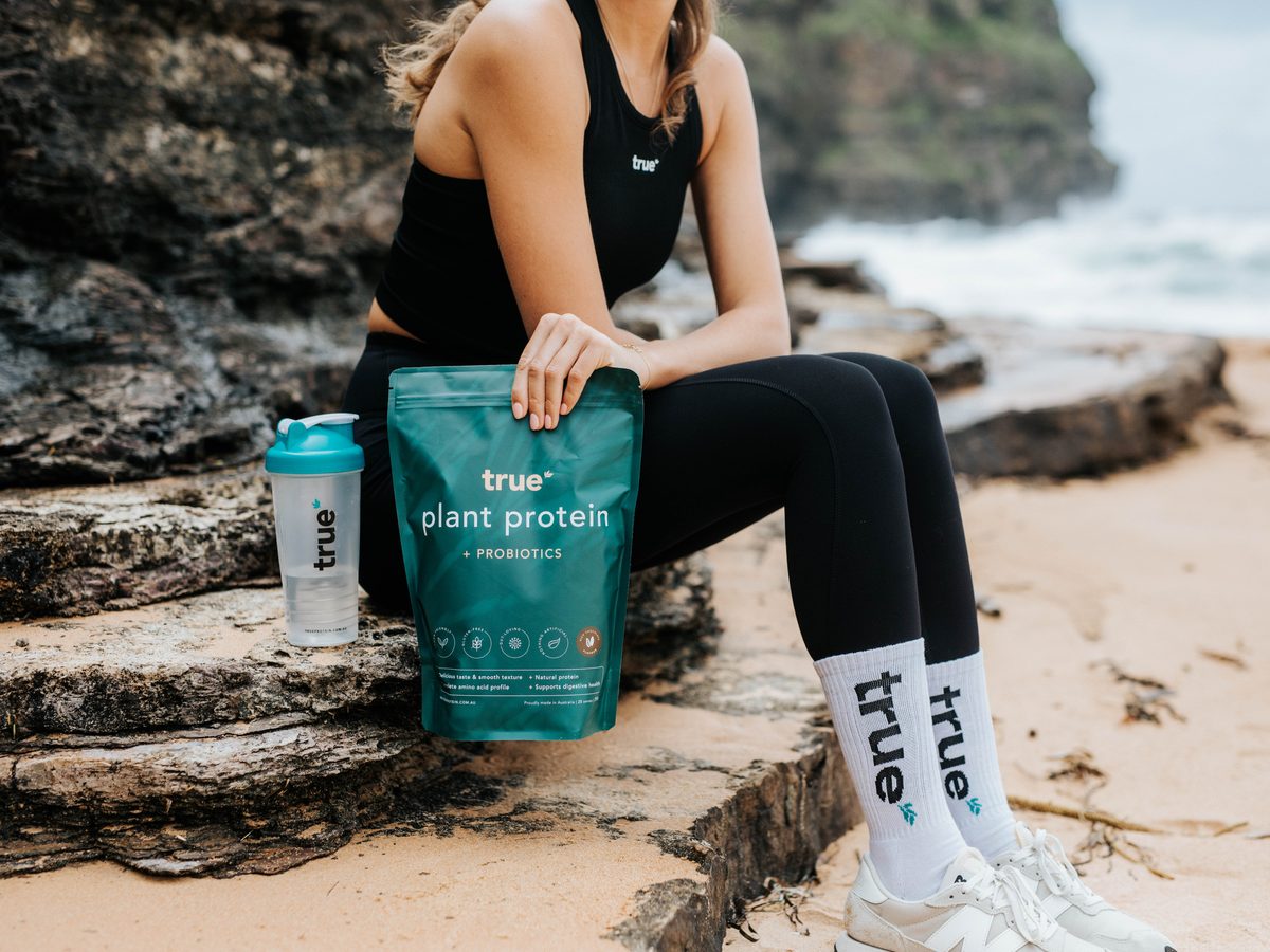 Woman in activewear holding a pouch of True Protein Plant Protein with Probiotics on a rocky Sydney beach