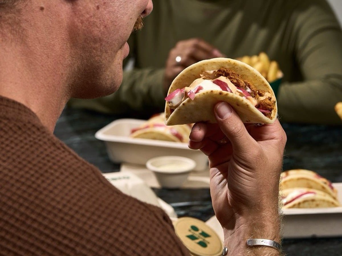 A man holding Zambrero's new Fat Taco in one hand at a table set with trays of tacos and fries