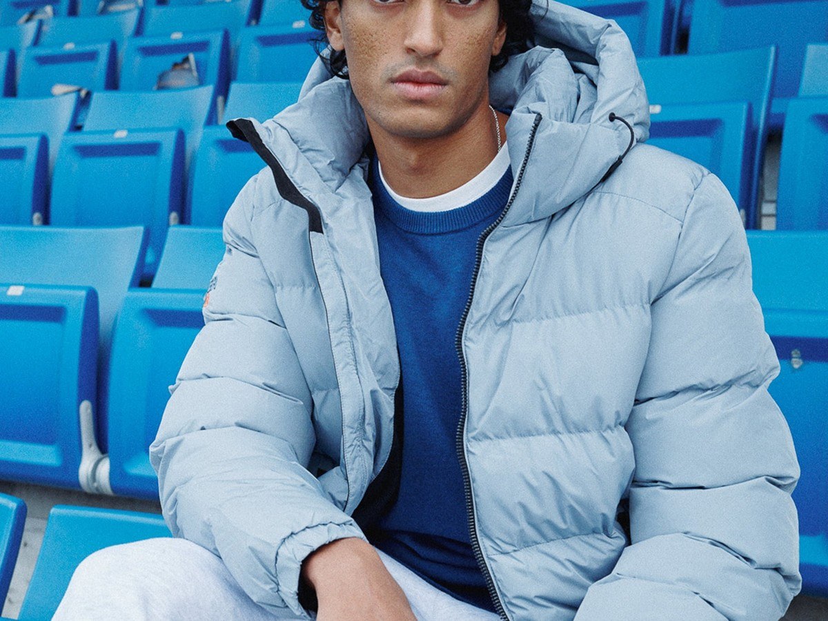 Man in a pale grey Superdry puffer jacket seated in blue stadium seats