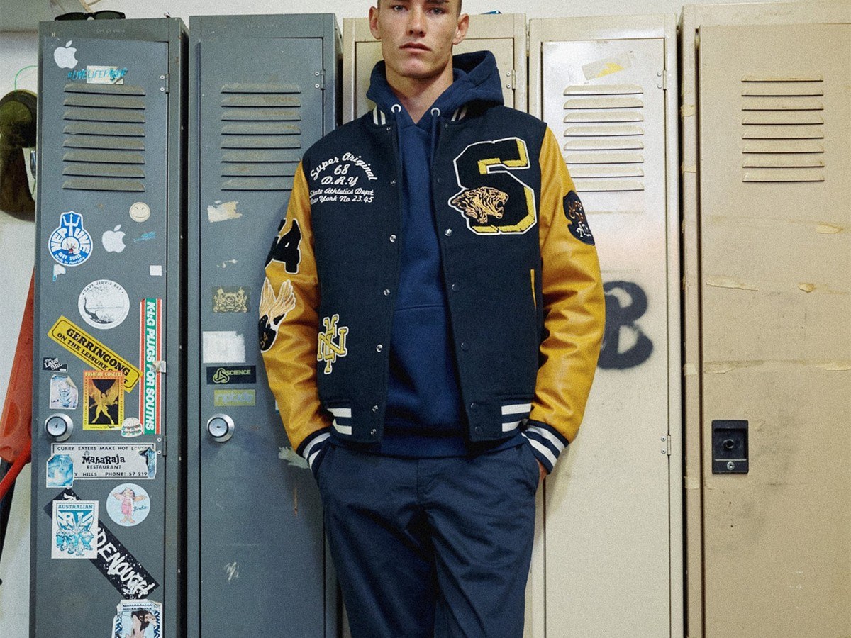 Man wearing a Superdry varsity jacket leaning against a row of lockers