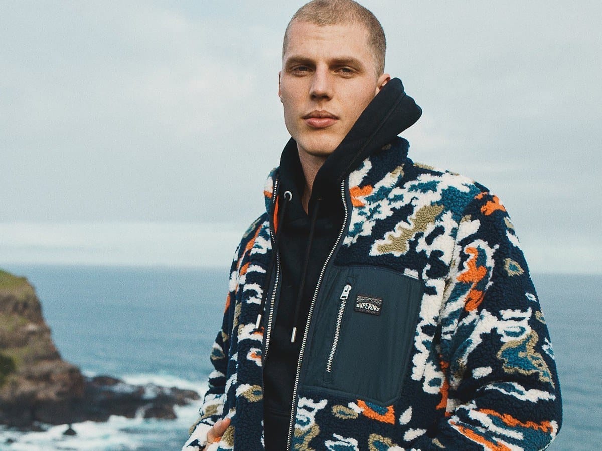 Man wearing the Superdry printed fleece zip-through jacket in lichen print on a coastal clifftop