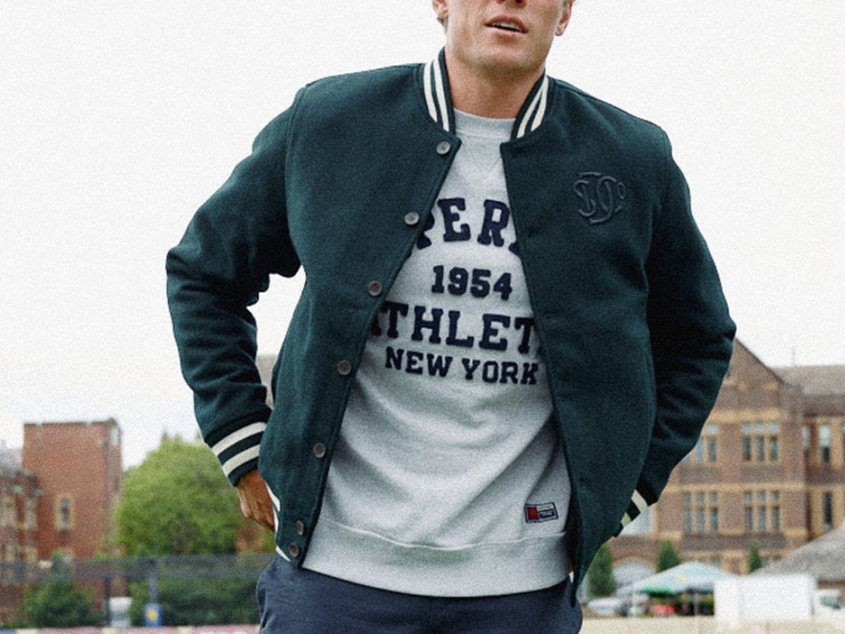 Man wearing the green Superdry varsity monogram wool bomber over a grey sweatshirt on a college green