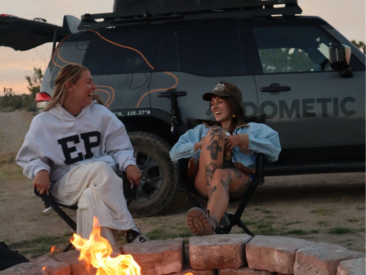 Two campers laughing by the fire beside a Dometic 4x4 at dusk