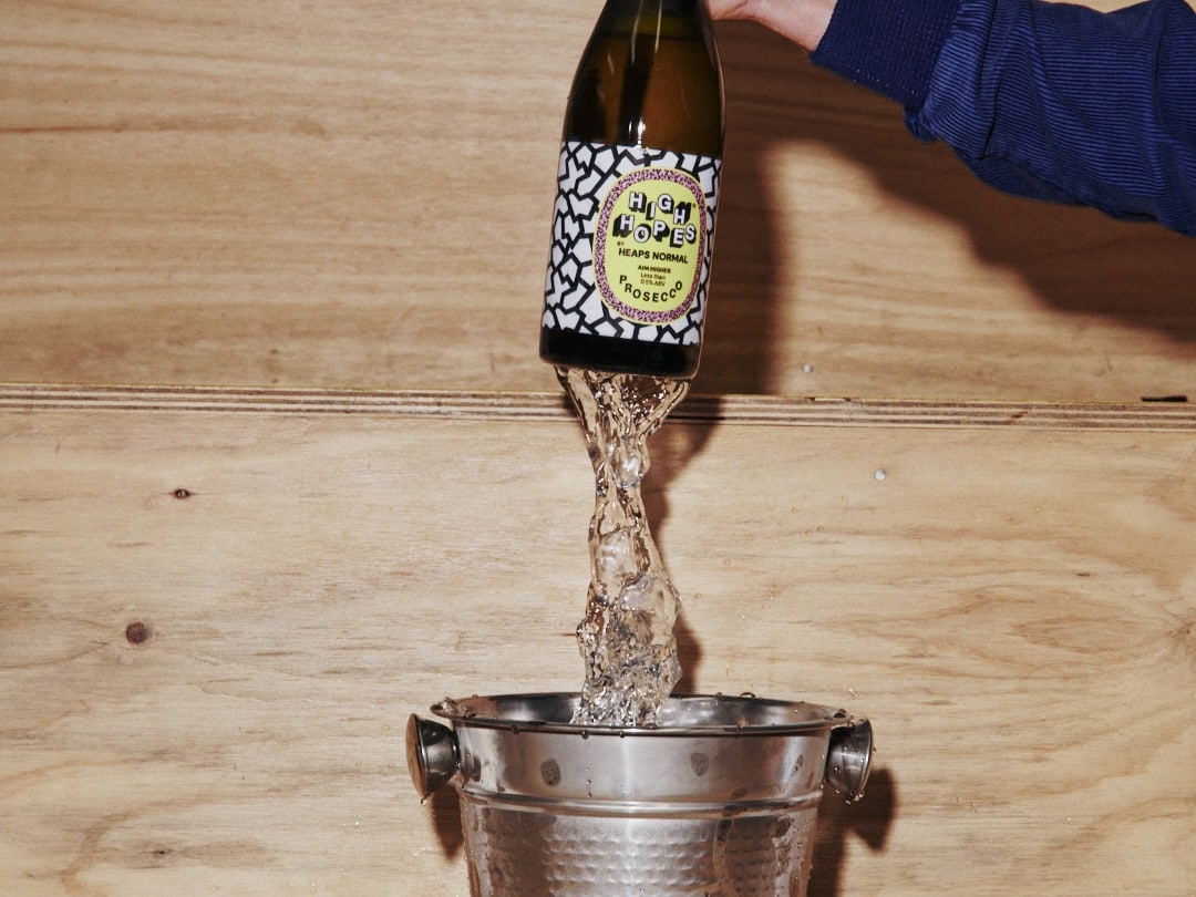 Hand pouring High Hopes prosecco into a silver ice bucket on a plywood bar