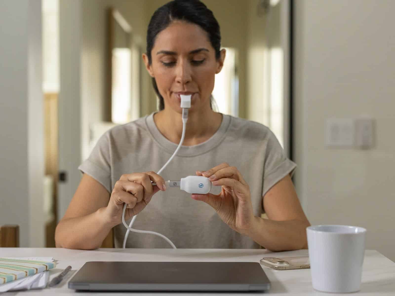 A woman running a daytime eXciteOSA session at her kitchen bench, with the mouthpiece connected to the handheld controller.