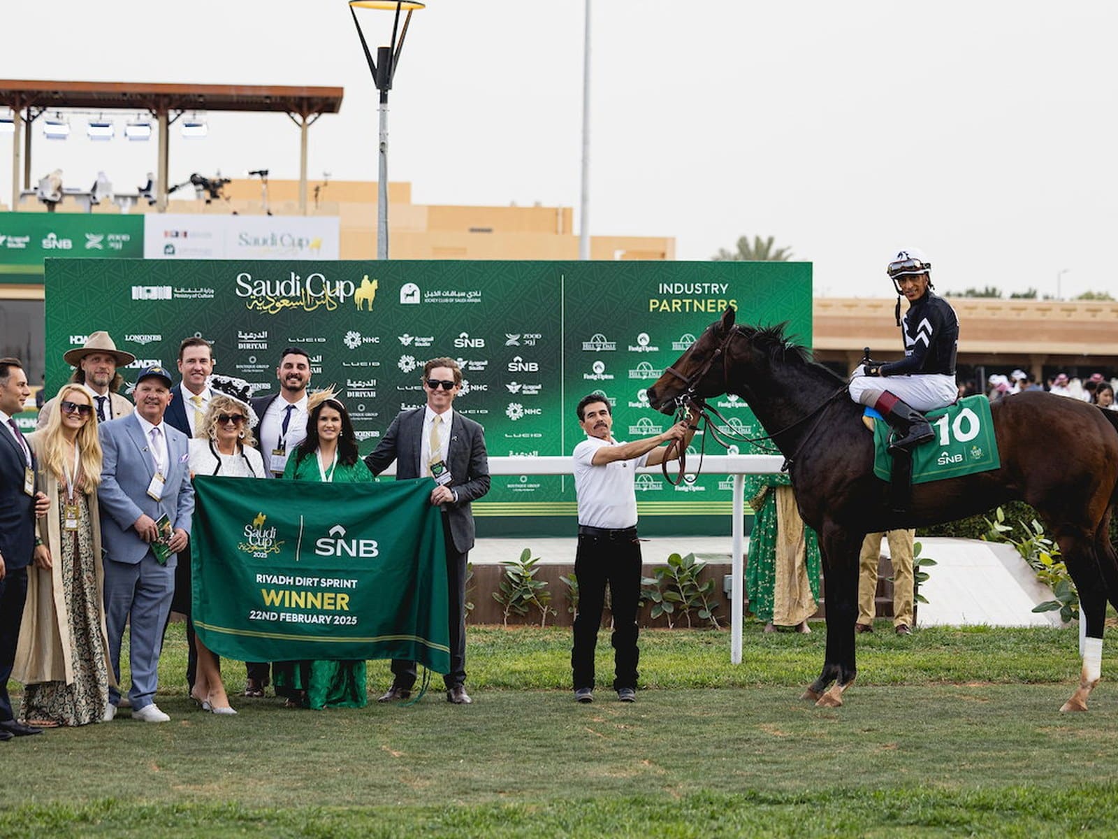 An owner group holds a Riyadh Dirt Sprint winner's banner on the turf beside the horse and jockey at the Saudi Cup meeting.