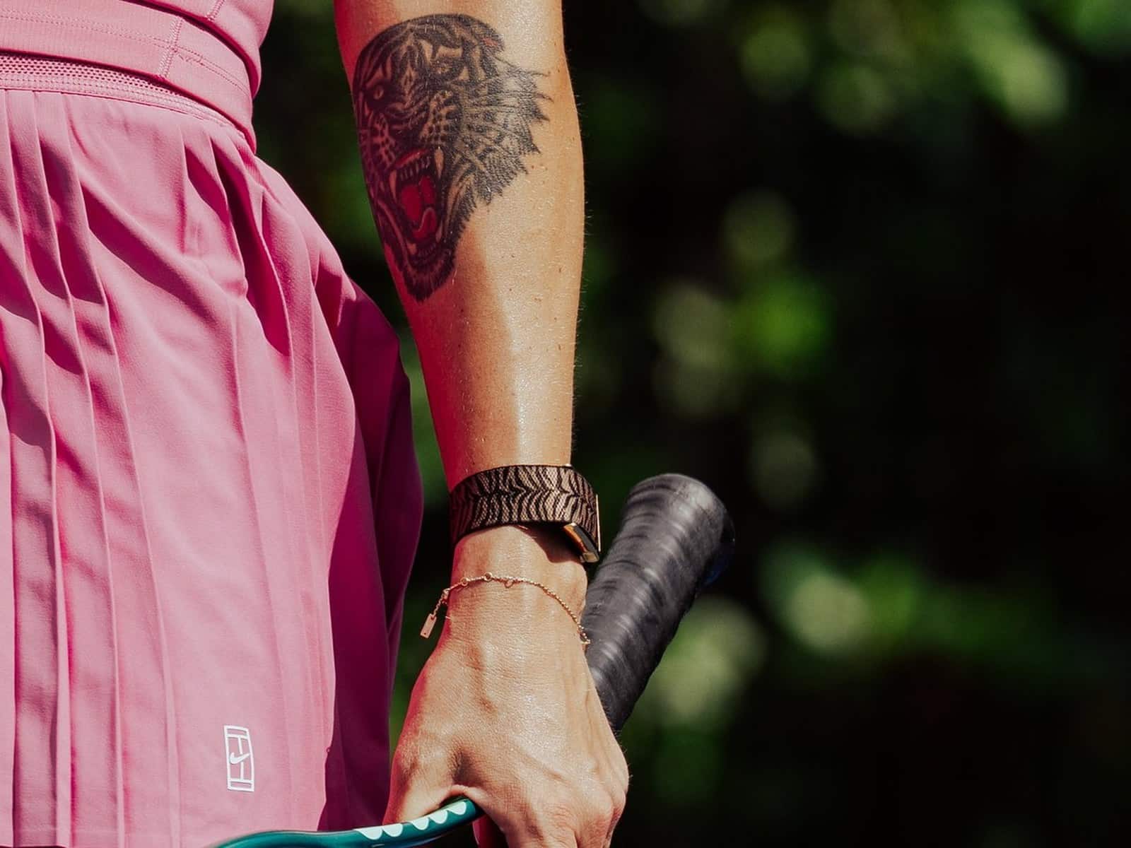 Close-up of Aryna Sabalenka's forearm showing her tiger head tattoo above the tiger-stripe WHOOP band on her wrist, holding a tennis racquet.