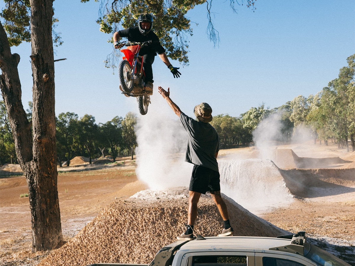 A dirt bike rider airborne mid-jump reaching out to high-five a person standing on the roof of a pickup truck at an off-road track.