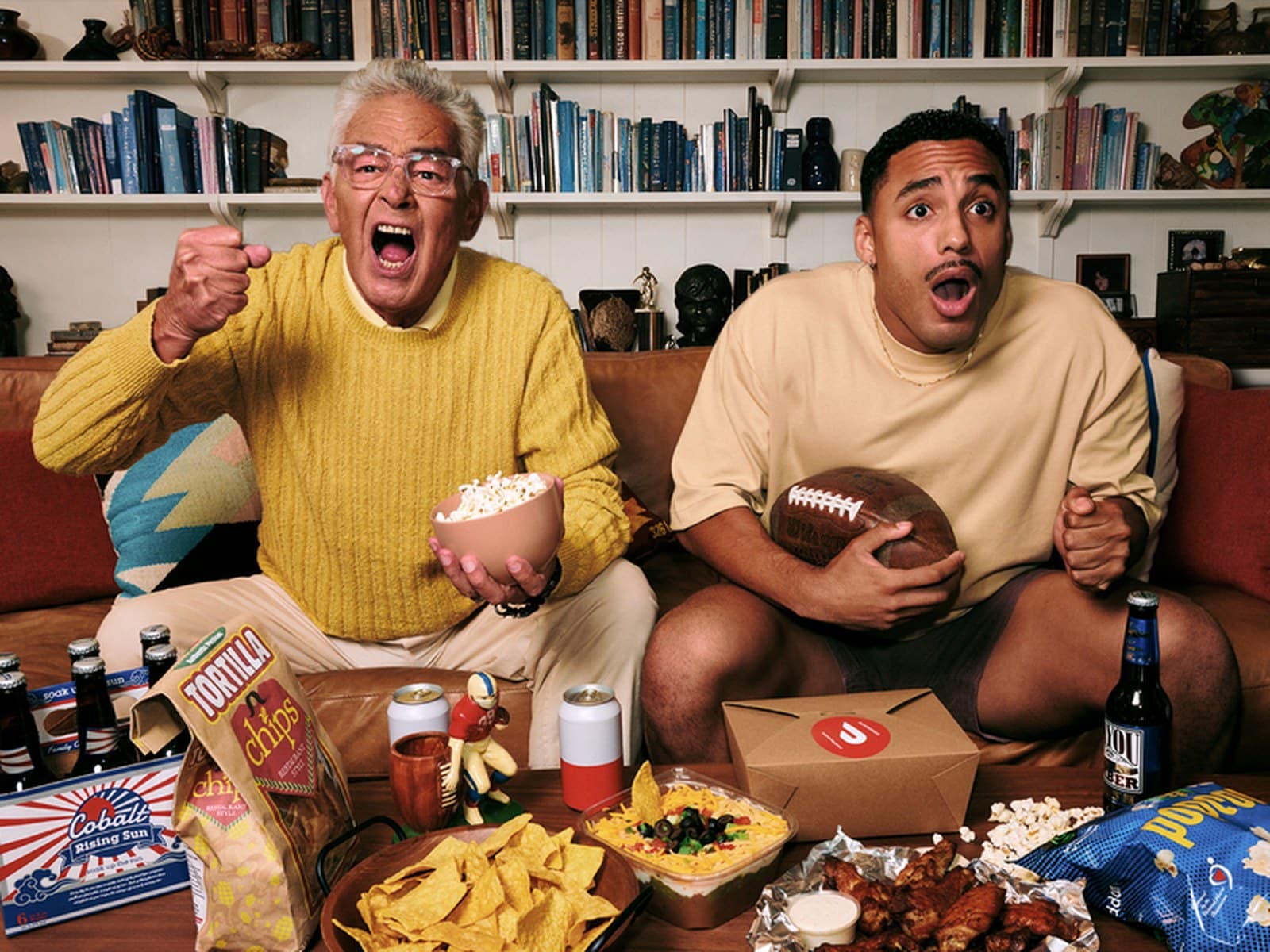 Fans watching a college football game on a couch with delivered food and drinks.