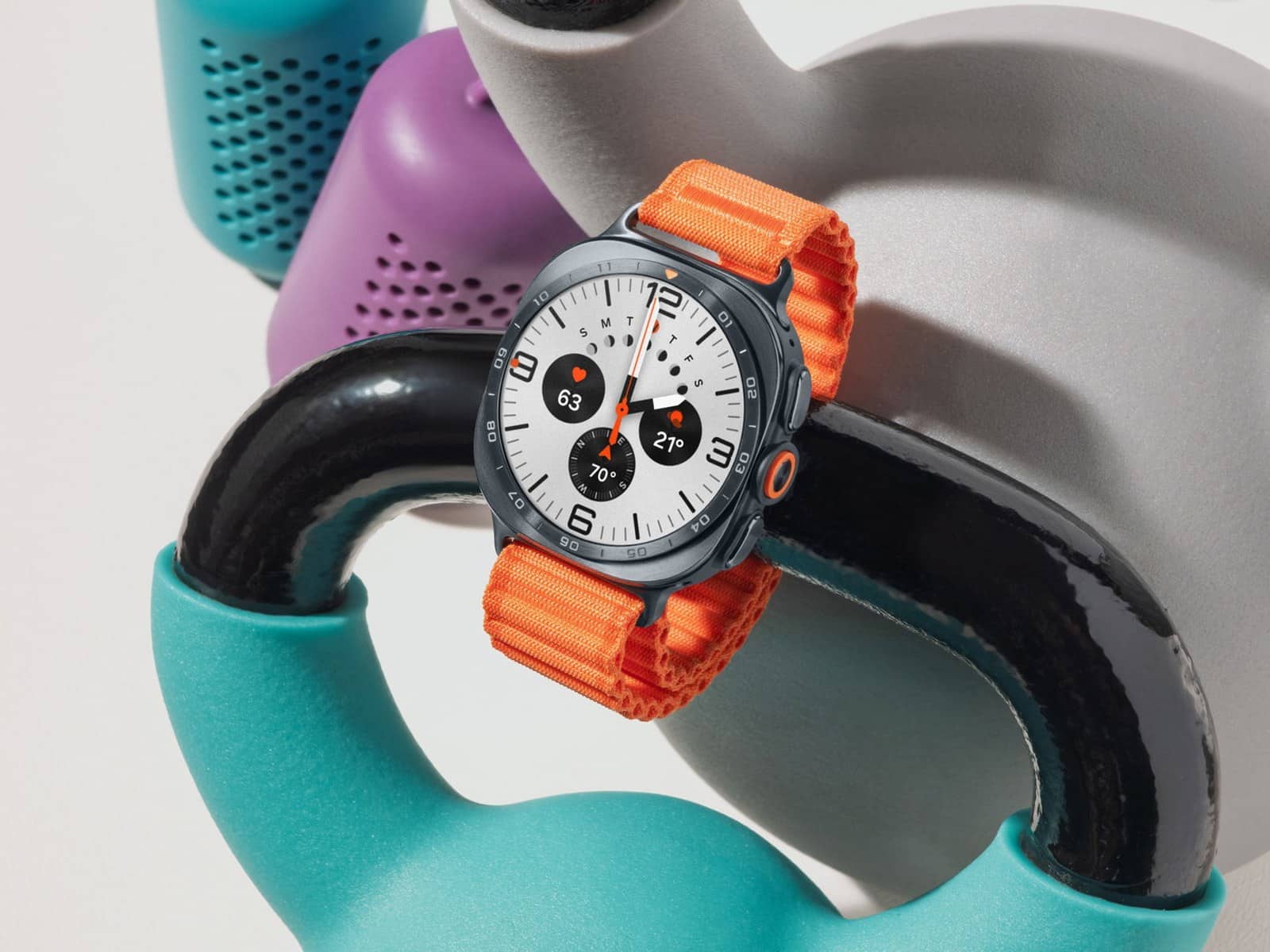 The Samsung Galaxy Watch8 Classic with a marked bezel ring and an orange woven band, resting on a set of coloured kettlebells.