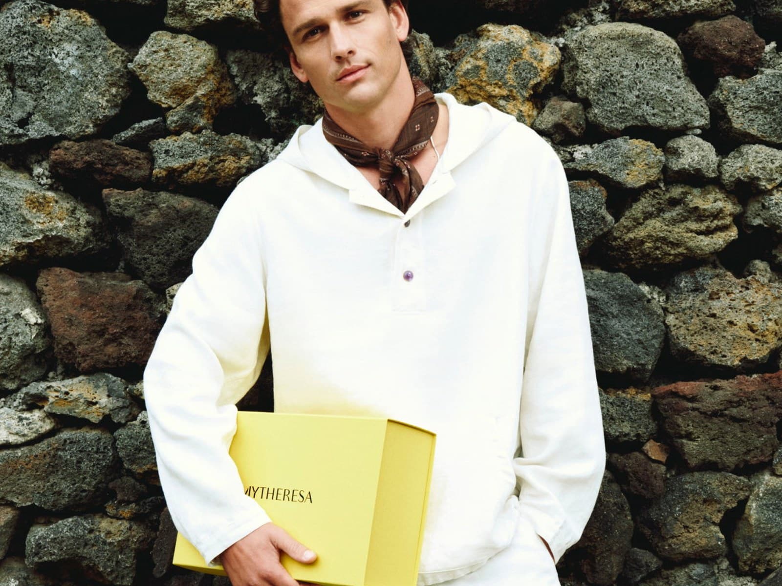 A model in a white hooded linen pullover and brown printed neckerchief against a stone wall.