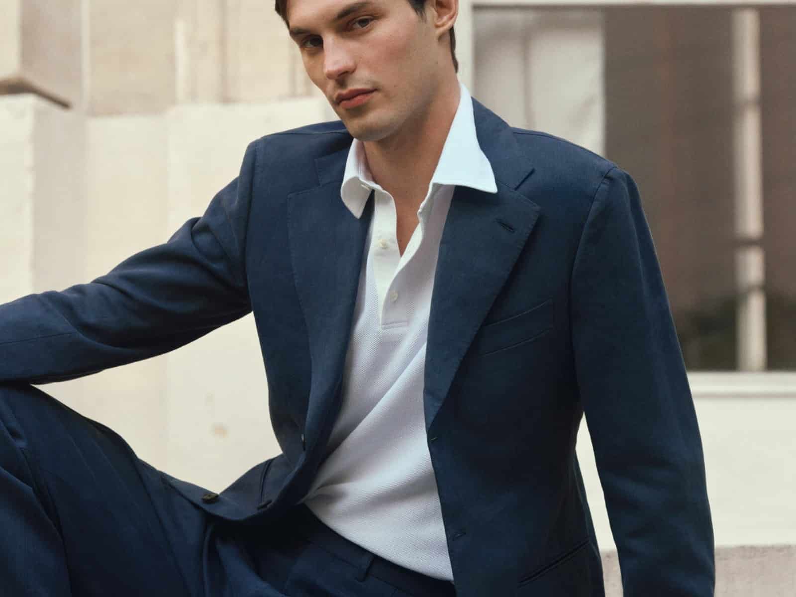 A model in a navy linen suit and white pique polo seated on stone steps.