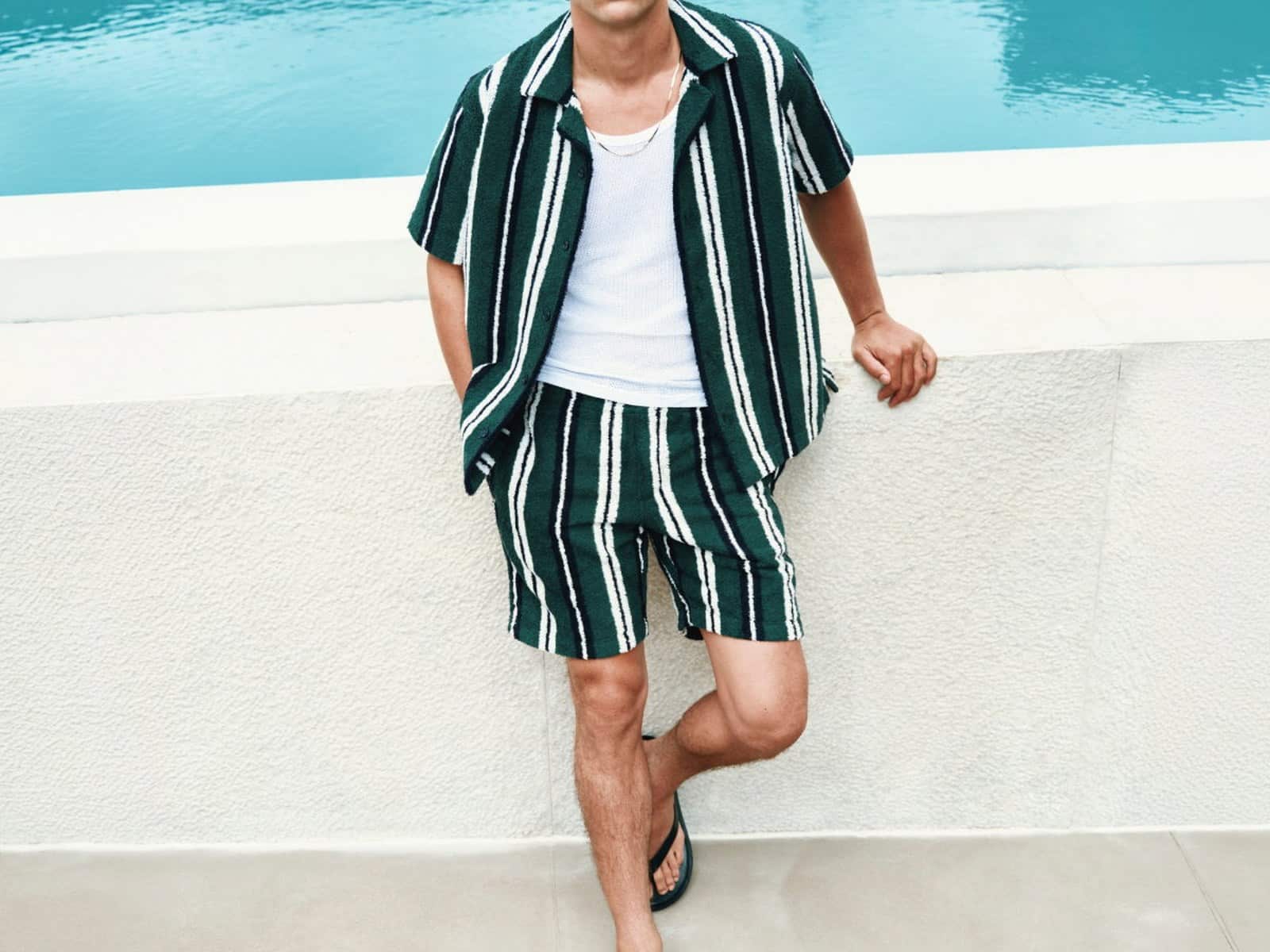 A model in a green and white striped terry camp-collar shirt and matching shorts beside a swimming pool.