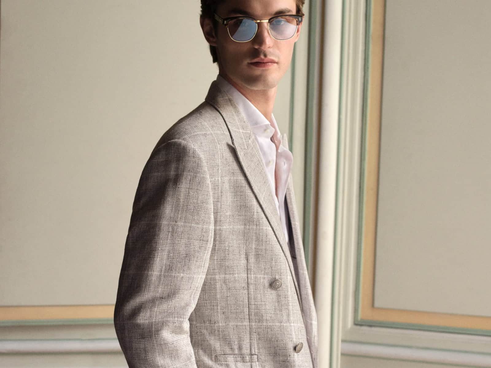 A model in a grey Prince of Wales check linen blazer and white shirt, standing beside panelled doors.