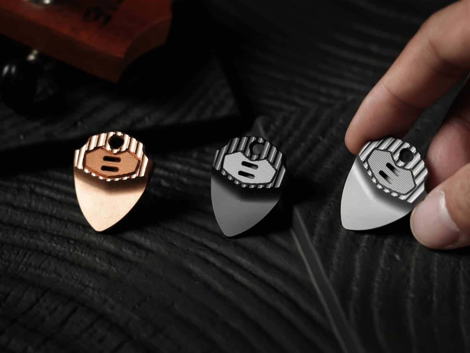 Three EchoTi picks in copper, dark grey and silver on black fabric, a hand reaching for one
