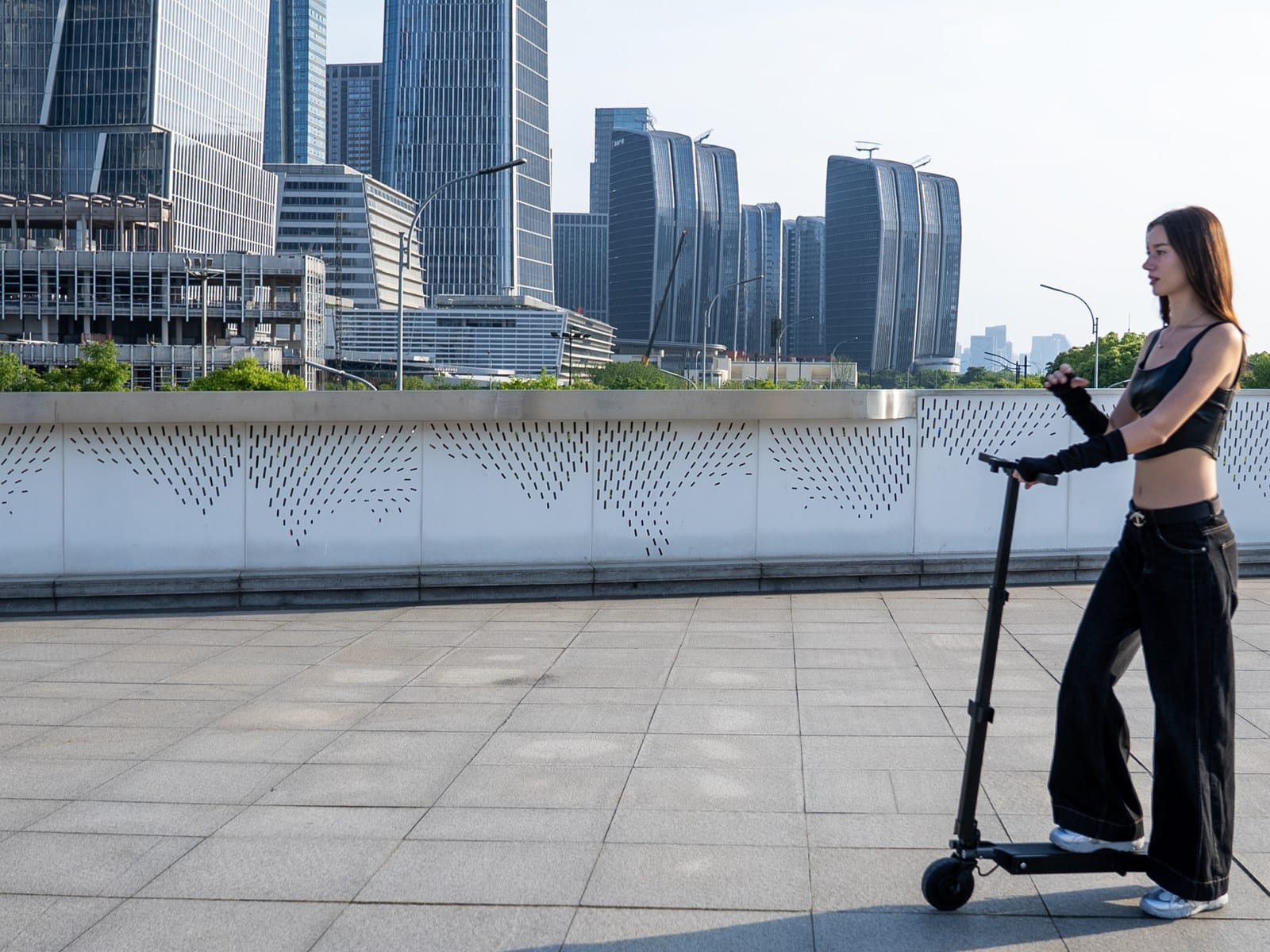 Woman riding the Blizwheel F0.5 electric scooter along a waterfront promenade with a city skyline behind her