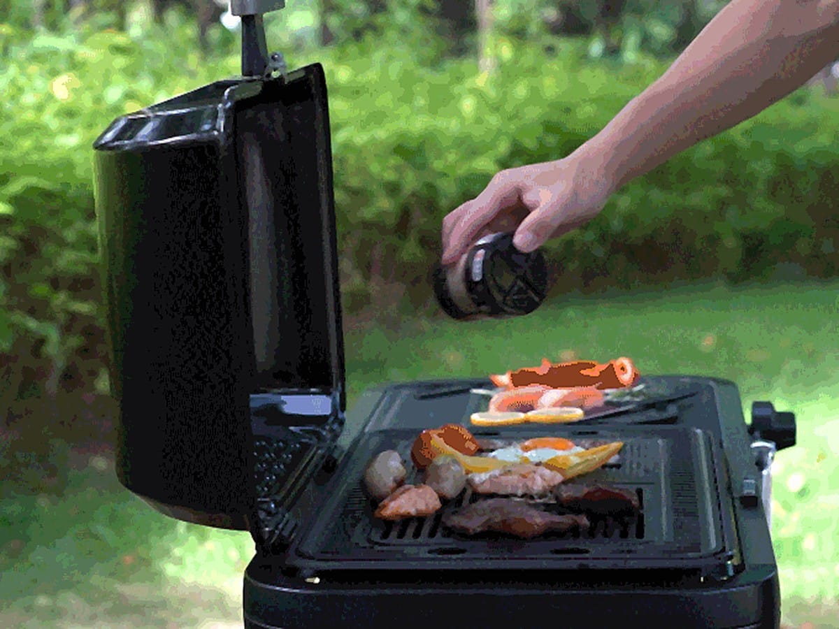 A hand reaching over the FYREL Fold & Go outdoor kitchen as prawns, eggs and vegetables cook on the grill plate, with the lid raised and greenery behind