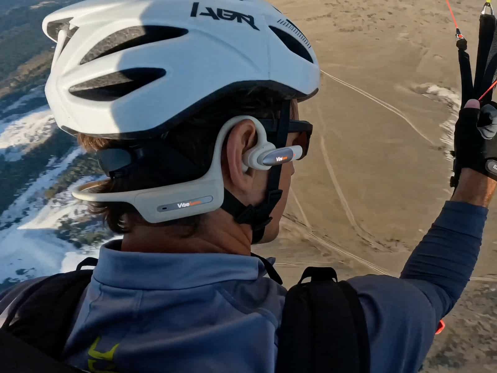 Paraglider pilot wearing a helmet and the MusicCam Pro headset, seen from behind with the camera arm visible at the ear and terrain far below