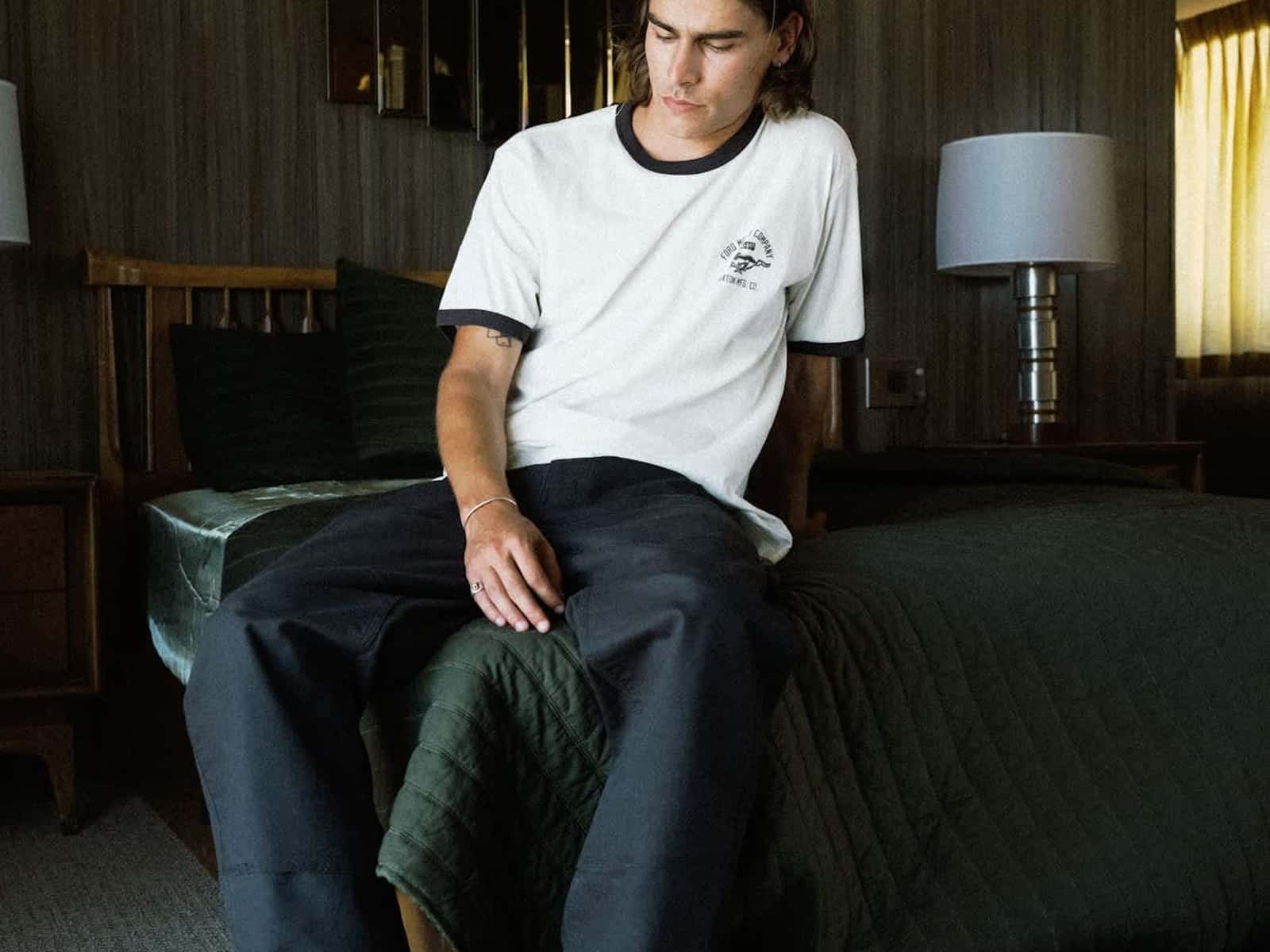 Model sitting on a motel bed wearing the Ford Mustang Ringer T-Shirt in Wimbledone White