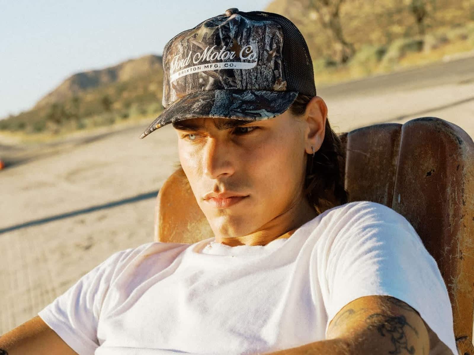 Model reclining in a desert chair wearing the Ford X Brixton Trucker Hat in Boxwood Green camo