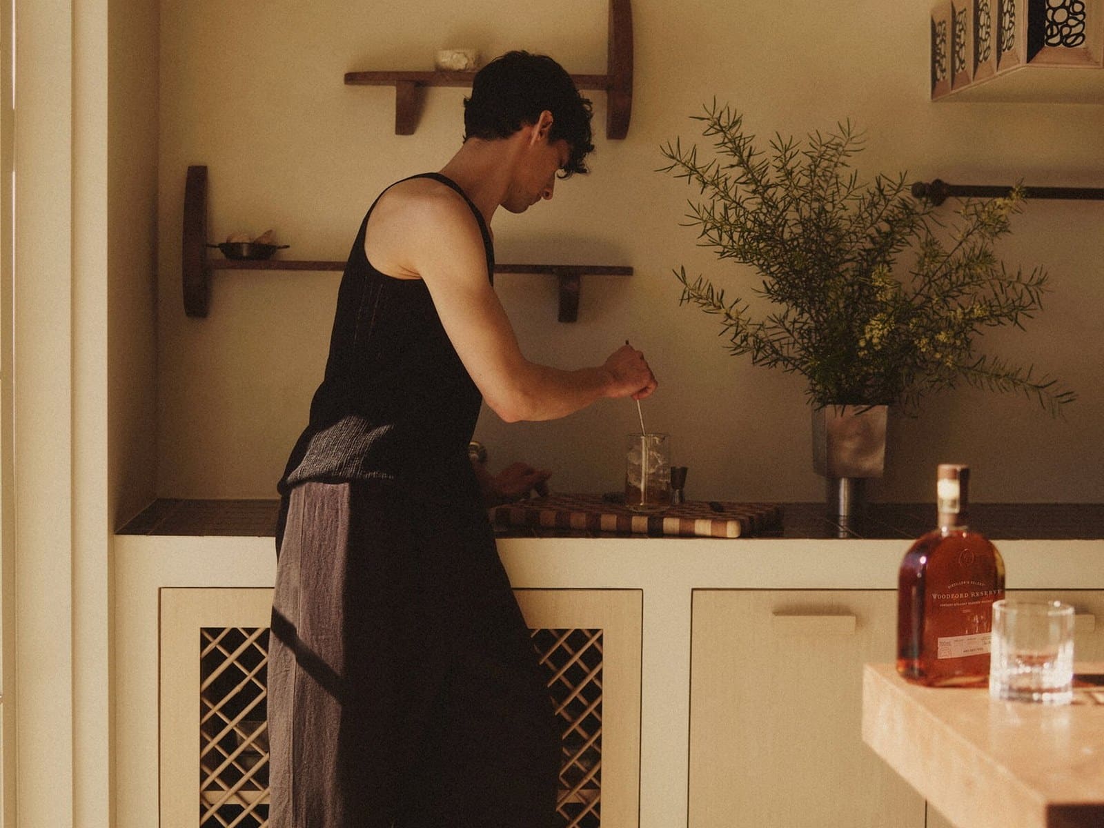 A man in a black singlet stirring a cocktail at a kitchen bench beside a bottle of Woodford Reserve.