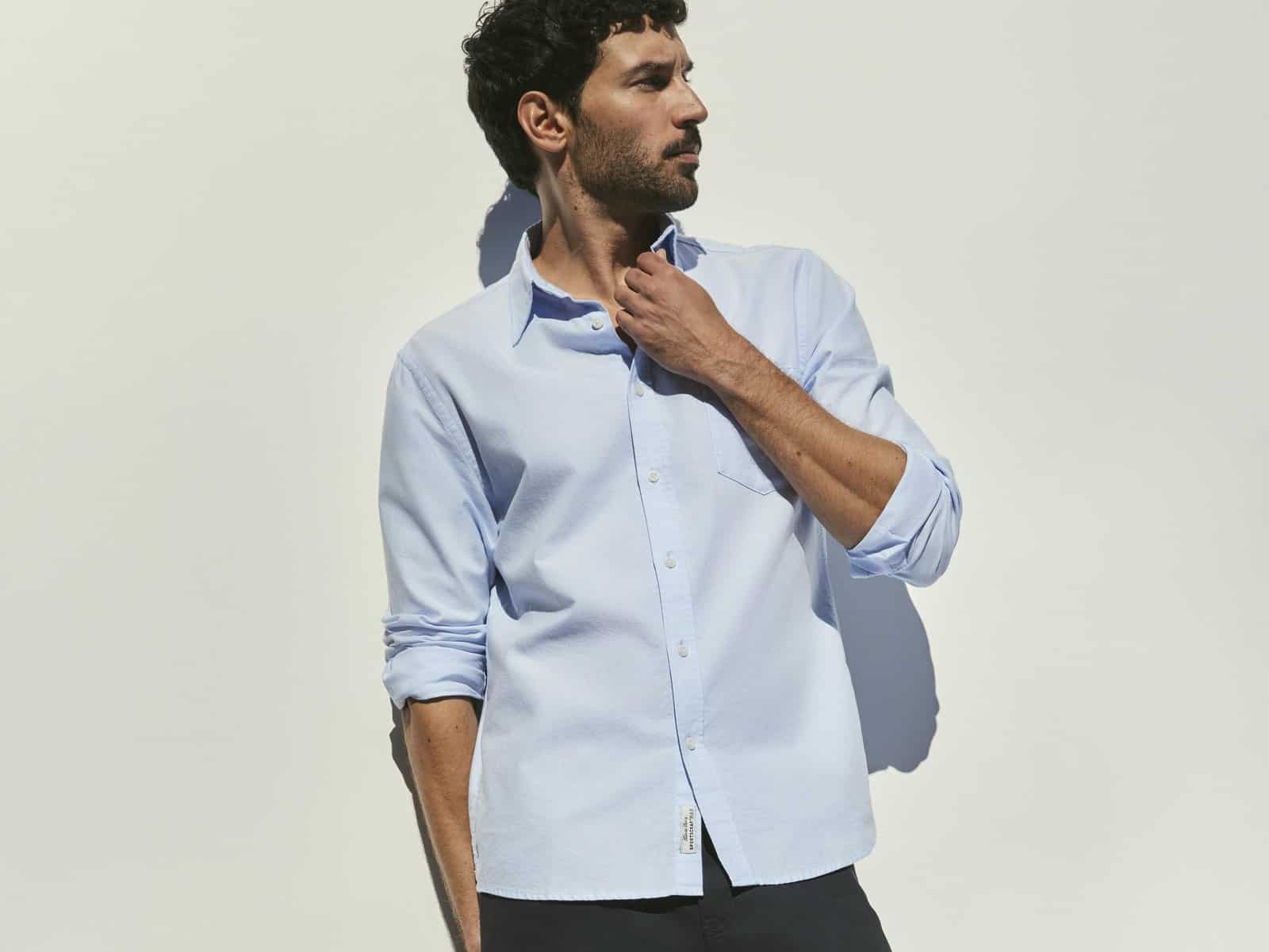 A man wearing the Sportscraft Long Sleeve Oxford Shirt in Light Blue with navy pants and a black leather satchel