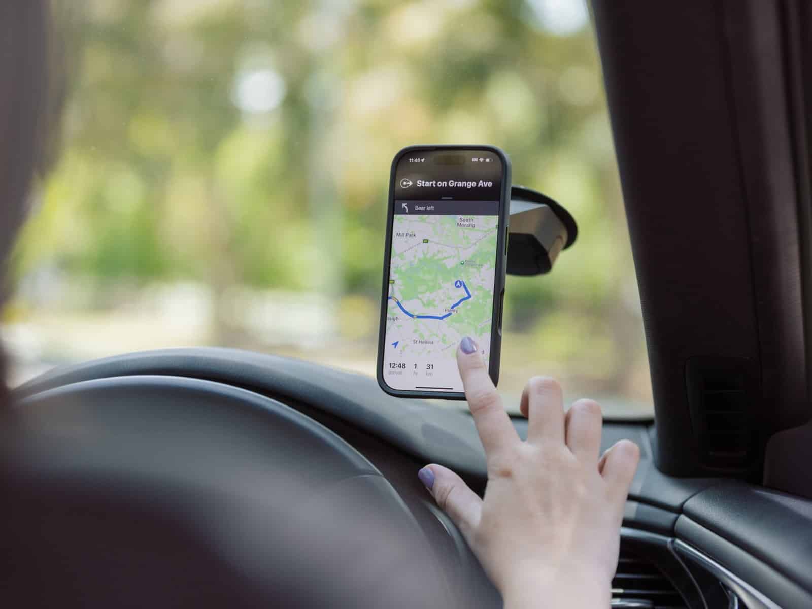 A driver reaching towards a phone in a Quad Lock case mounted on a car windscreen displaying a maps navigation route