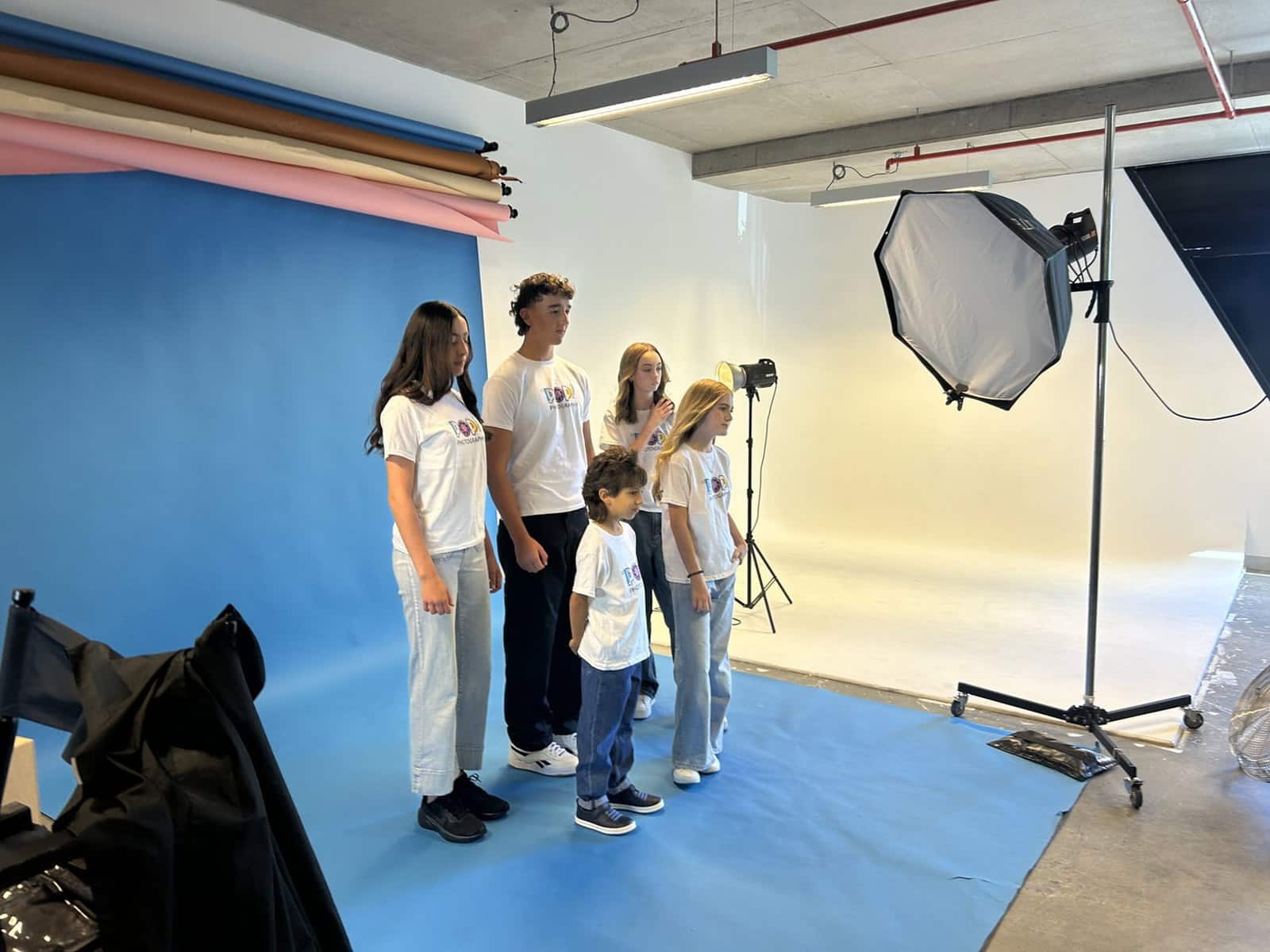 Five young models in matching white T-shirts stand on a blue paper backdrop in a POP Photography studio while a softbox light is positioned beside them