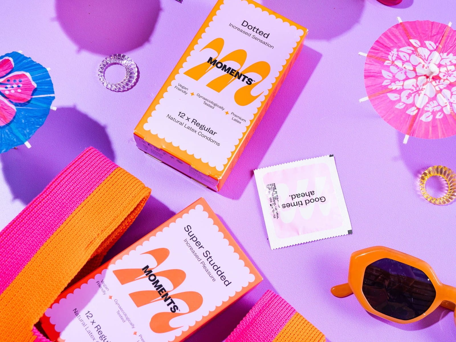 Flat lay of Moments condom boxes and sachets among cocktail umbrellas on a lilac background