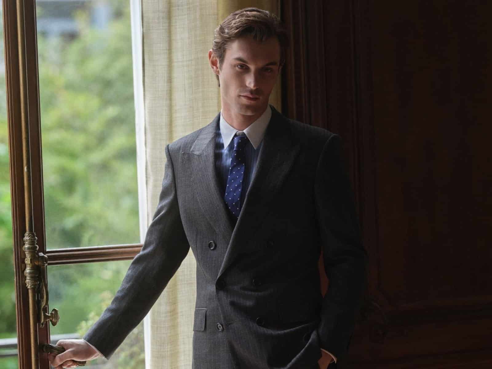 A model in a charcoal pinstripe double-breasted suit, white shirt and navy polka-dot tie, standing beside a tall window with a brass handle.