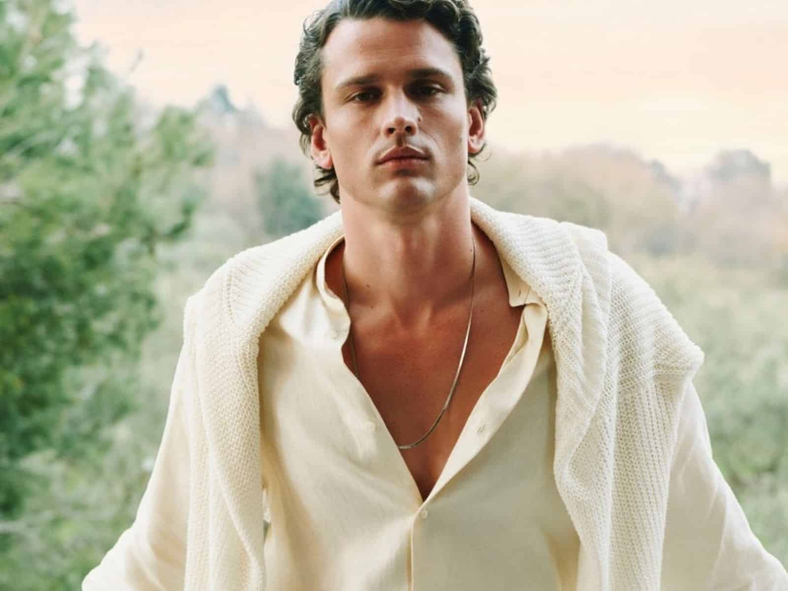 A model in a cream open-collar shirt with a cream knit draped over the shoulders, standing in front of olive trees at dusk.