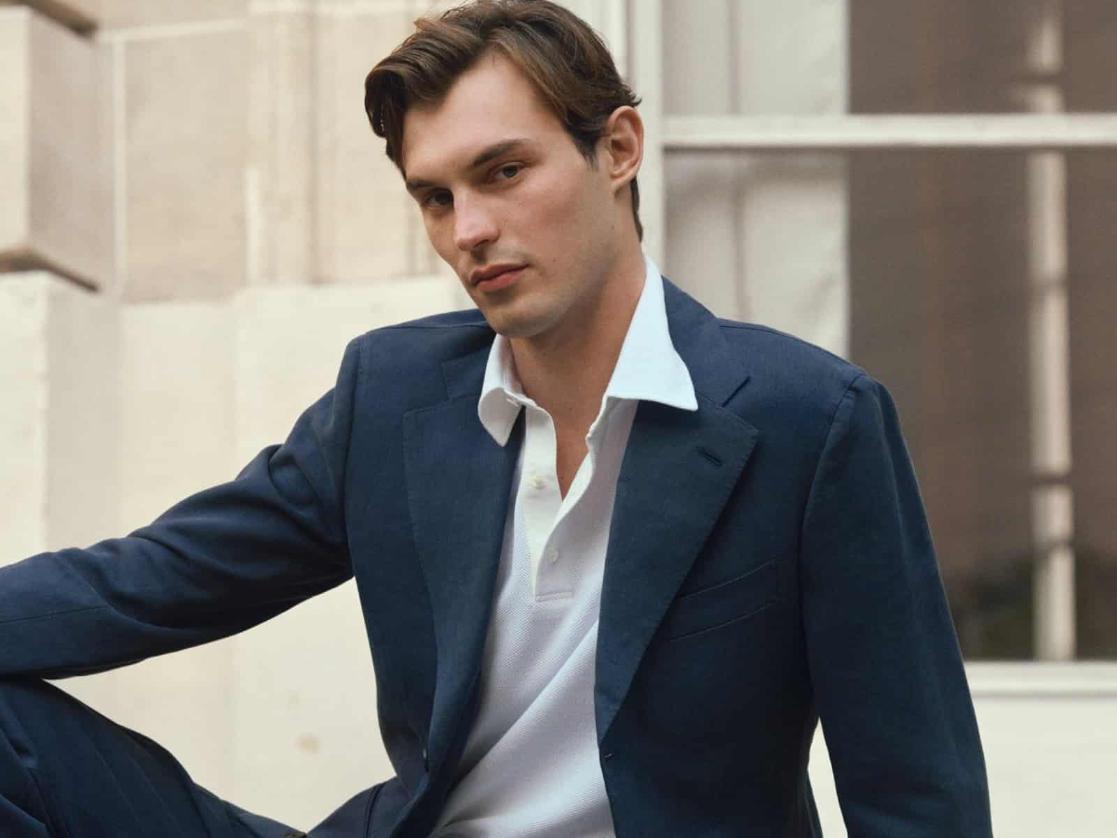 A model in a navy suit and white piqué polo, seated on stone steps outside a stone building.