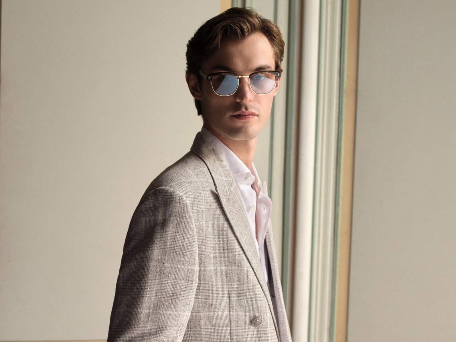 A model in a grey Prince of Wales check blazer and white open-collar shirt, standing beside panelled doors.