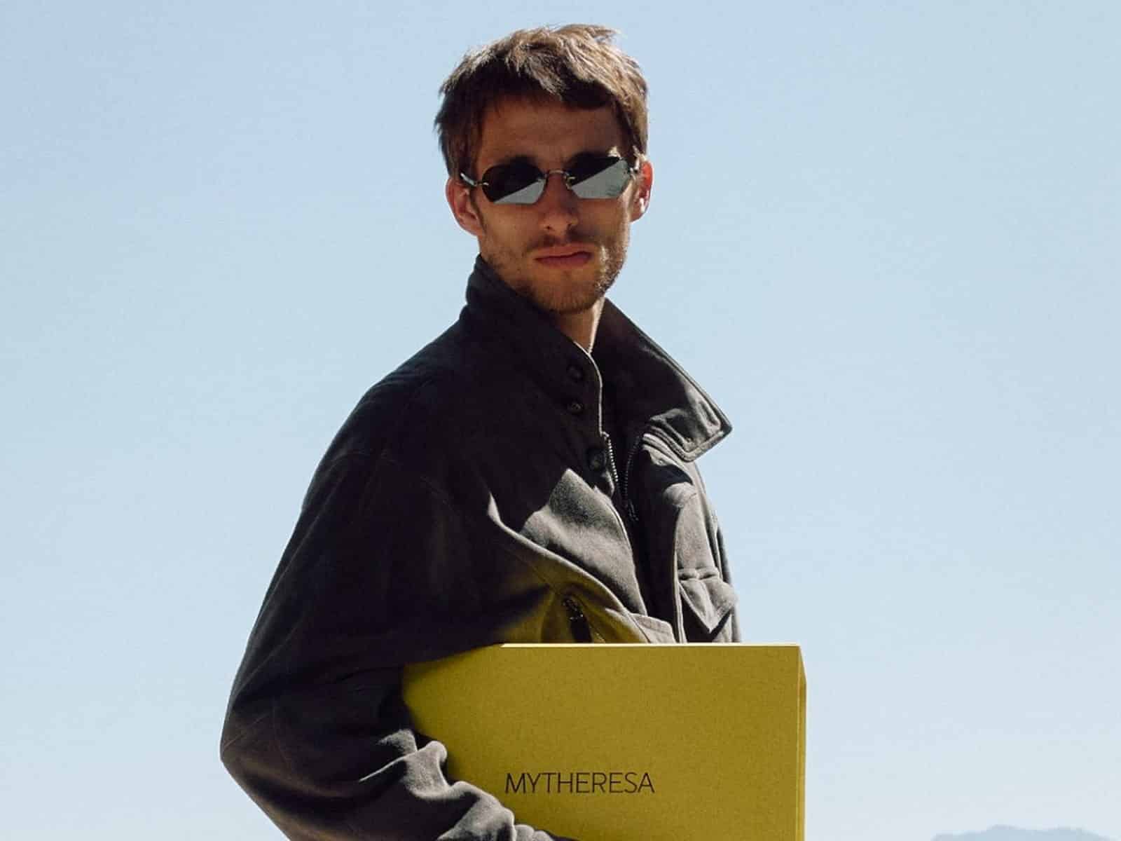 A model in sunglasses and a dark suede field jacket holding a yellow Mytheresa box, with mountains behind him.