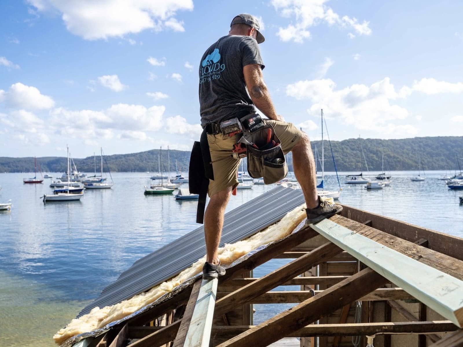 Tommy Alsop in work shorts and a tool belt standing on exposed roof framing above a bay full of moored boats