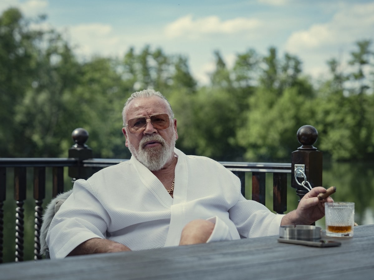 "Older man in white robe sits outdoors by a wooden table, holding a cigar and drink, with greenery and a lake in the background, under a partly cloudy sky."
