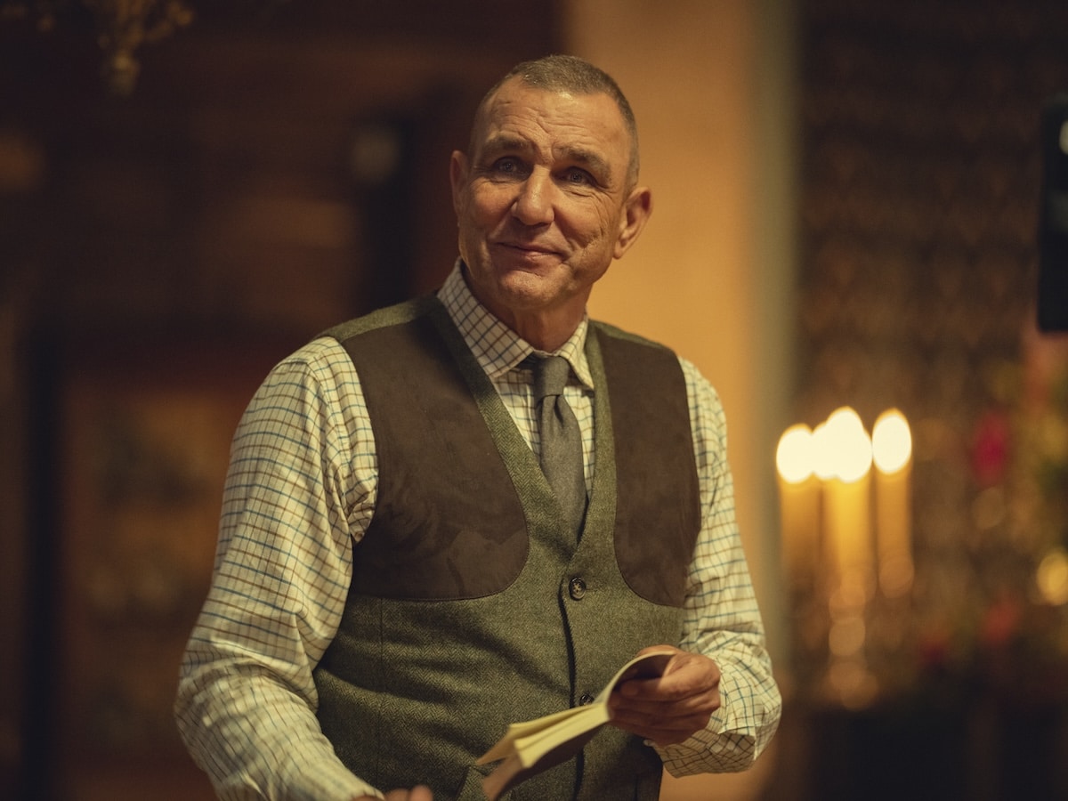 "Middle-aged man in a checkered shirt and vest holding a book, standing indoors with warm lighting, blurred background features candles and wooden decor, creating a cozy, intimate atmosphere."