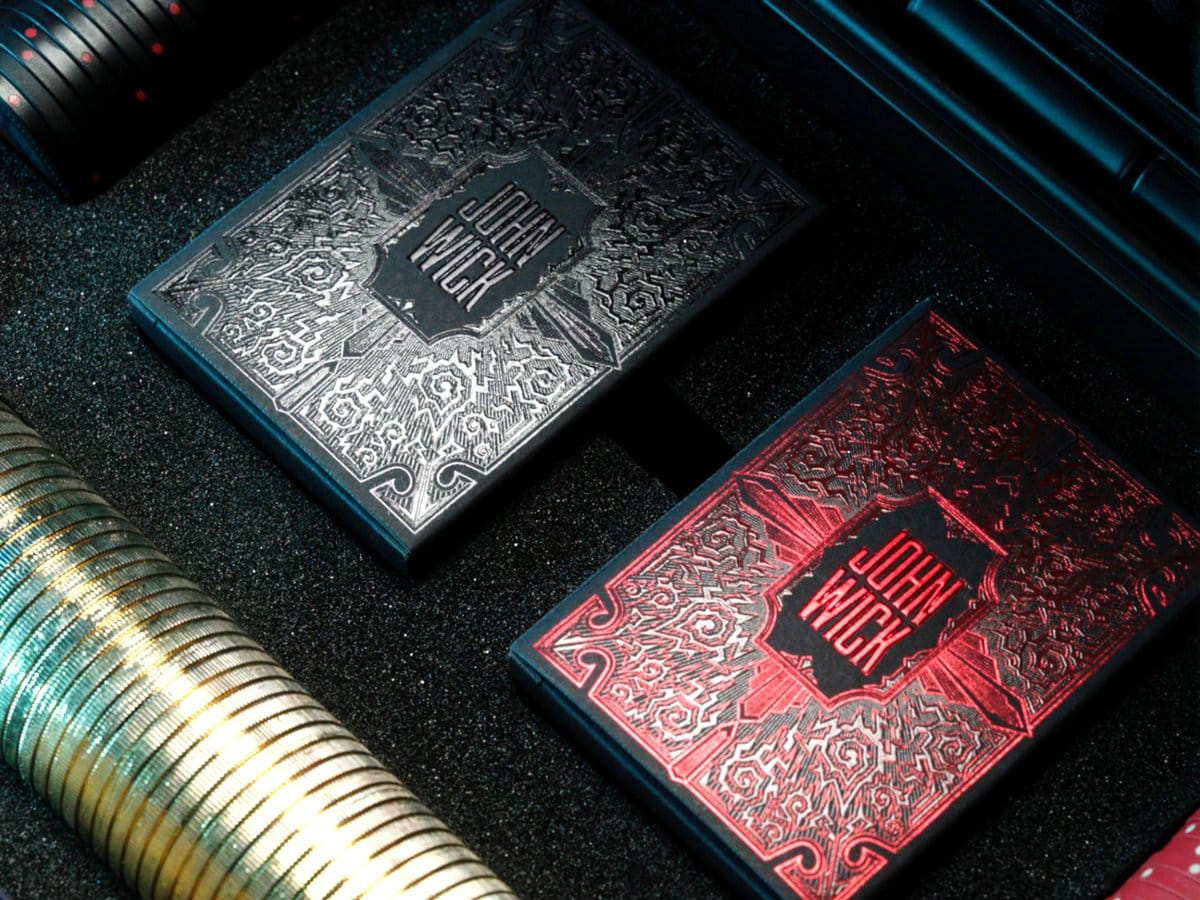The two John Wick collector's card decks, one silver and black and one red and black, beside a stack of gold coins