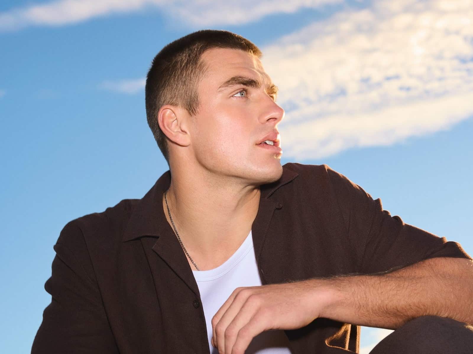A man in a dark short sleeve shirt over a white tee, crouching against a bright blue sky