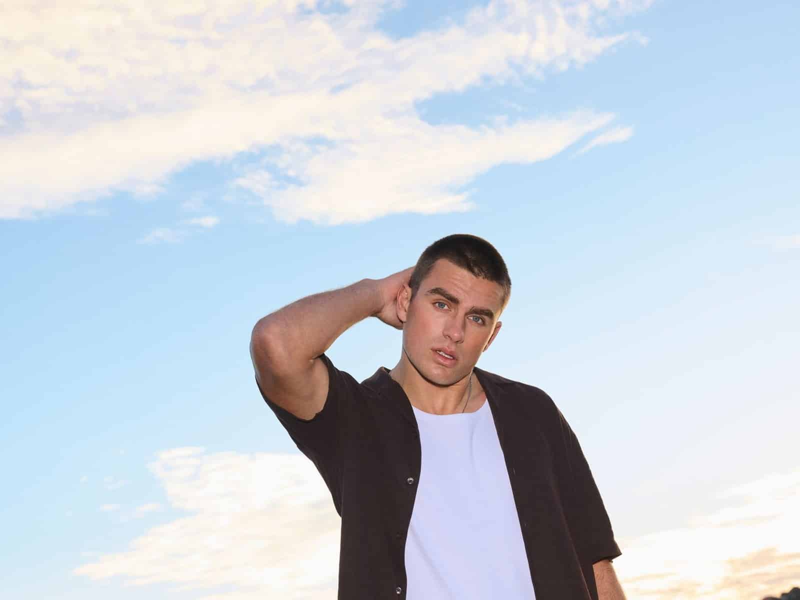 A man in an open dark short sleeve shirt and white tee standing by the harbour at sunset, one hand behind his head