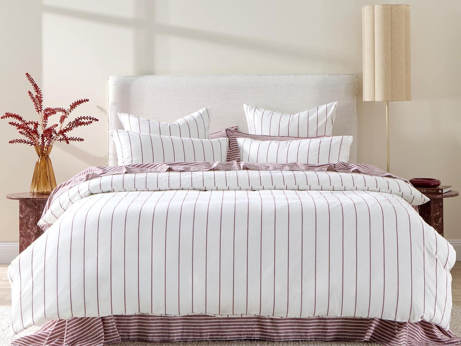 A bed made up with a white and burgundy pinstripe quilt cover set and matching striped valance, with a dried flower arrangement on a side table.