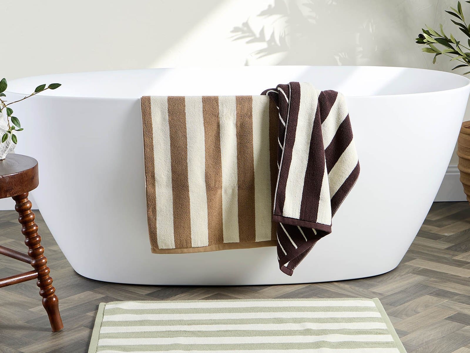 Tan and chocolate striped towels hung over a freestanding white bath, with a sage and cream striped bath mat on a herringbone timber floor.
