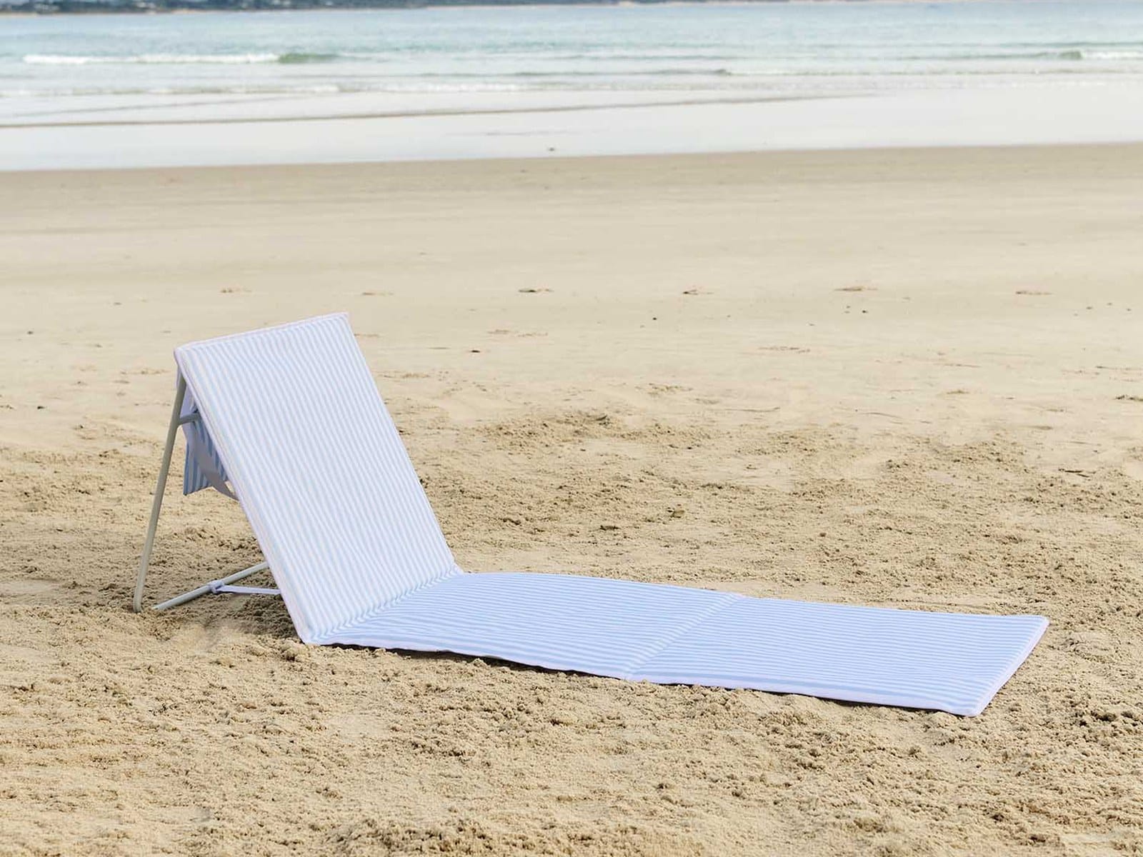 A sky blue and white striped reclining beach lounger set up on the sand with the ocean and distant hills behind.
