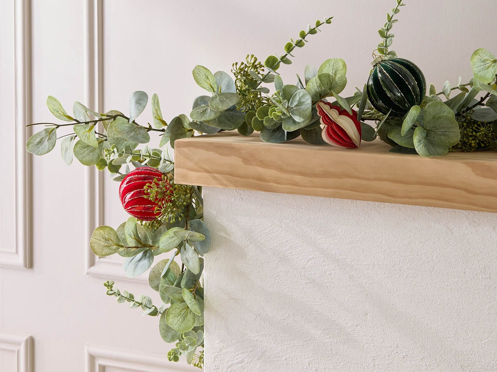A faux eucalyptus Christmas garland with red and green baubles draped along a timber mantelpiece against a white panelled wall.
