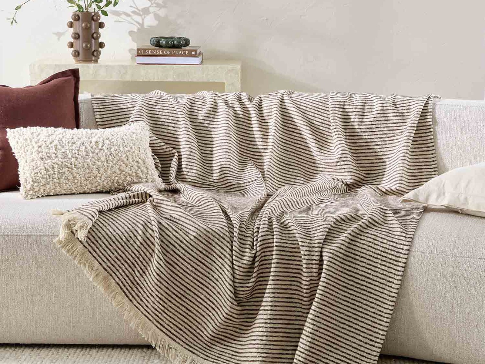 A cream and brown pinstriped fringed throw draped over a cream sofa with a textured boucle cushion and a rust linen cushion.