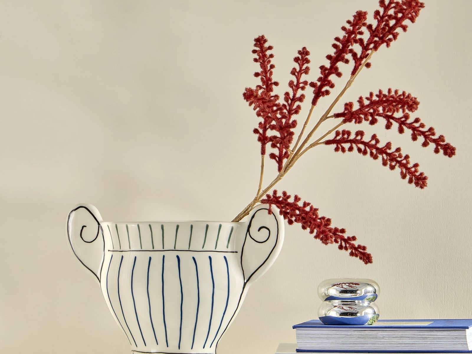 A white ceramic urn vase with hand-painted blue stripes and scrolled handles, holding red dried stems, on a stack of books beside a small glass ornament.
